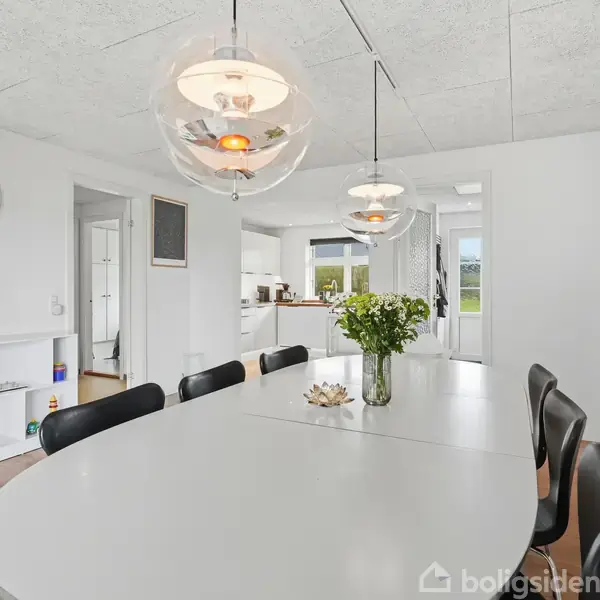 En spisebord med sorte stole står i en lys spisestue. Over bordet hænger tre lamper. I baggrunden er der en åben dør mod køkkenet og en reol med legetøj.