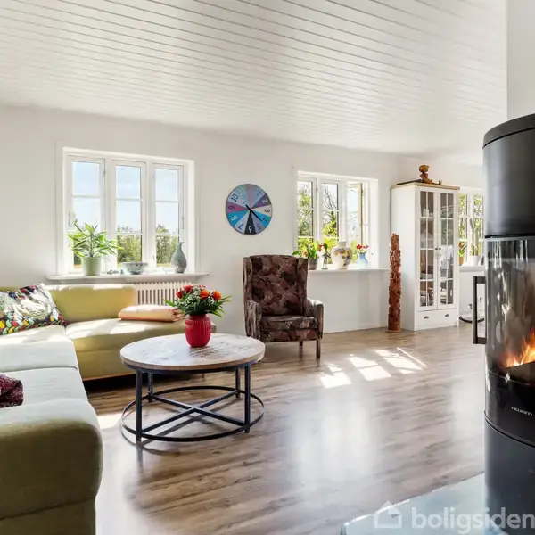 En sort brændeovn med ild brænder i et lyst stuerum. Rummet har en grøn sofa, et rundt sofabord, farverige puder og store vinduer, der lukker sollys ind.