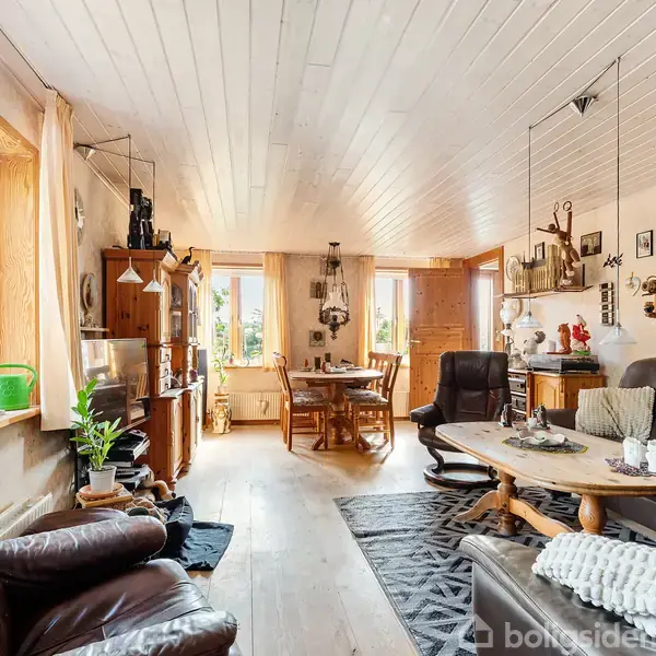 Stue med lænestole og sofa omkring et sofabord, dekoreret med tæppe og små pyntegenstande. Bagerst ses et spisebord nær store vinduer, der slipper lys ind.