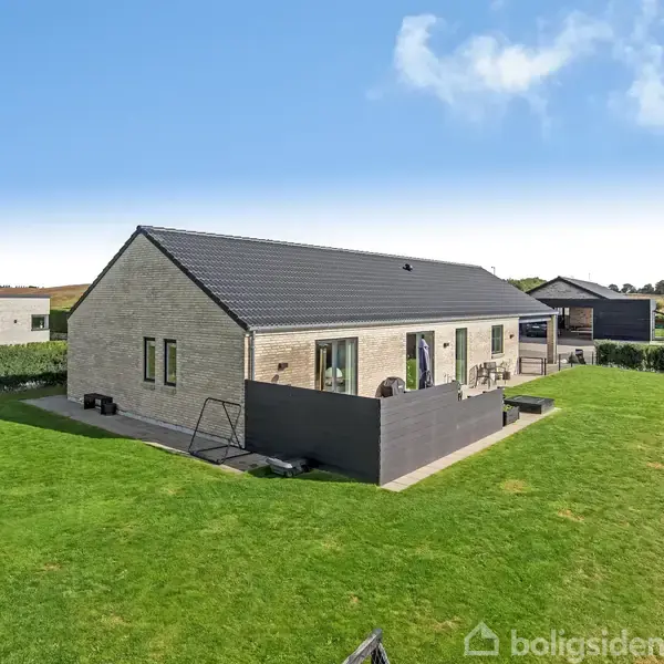 Et moderne hus står stille på en velplejet græsplæne omgivet af lave hække. Skyfri himmel bag huset, hvilket skaber en lys og åben atmosfære.