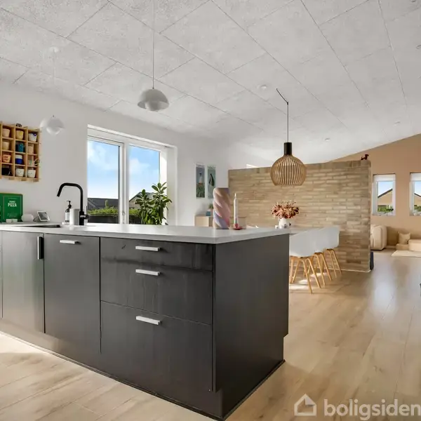 Køkkenbord med sort skab står i midten, omgivet af lyse trægulve og åbne vinduer; i baggrunden ses en spisestue med spisebord, stole og planter.
