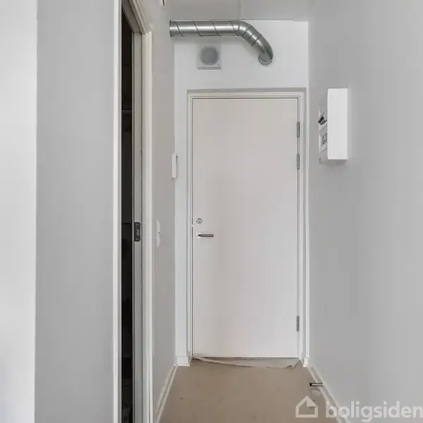 En hvid dør i en smal, grå gang ved siden af et moderne køkkenområde med en ovn og flasker. Ventilationsrør ses over døren.