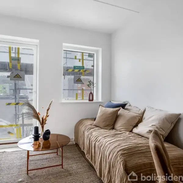 Sofa samlede puder i lys stue, placeret ved to vinduer med støvede rosetter. Rundt bord med tørrede blomster i vase. Lyse vægge og gulvtæppe skaber behagelig atmosfære.