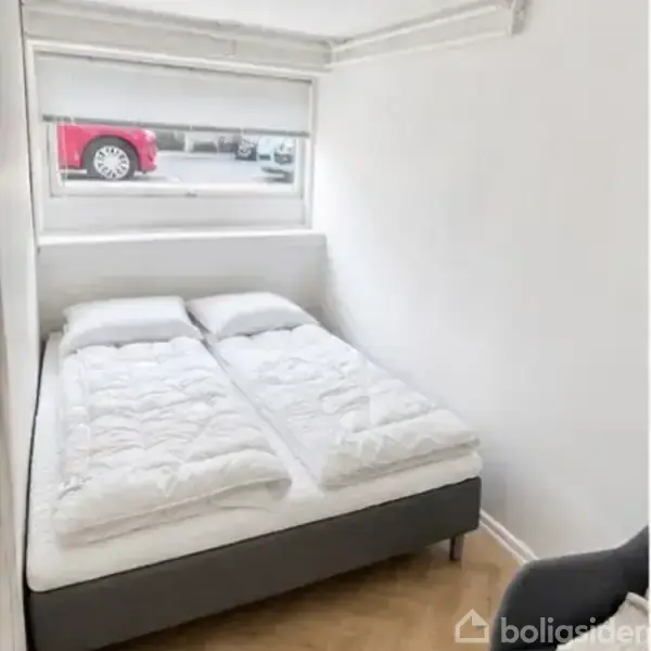 En dobbeltseng med to hvide dyner står i et lille rum. Gennem et vindue ser man parkerede biler udenfor. Rummet har hvide vægge og minimalistisk indretning.