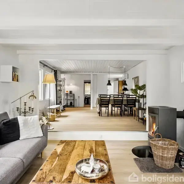 Grå sofa står ved siden af et træbord. I baggrunden ses en spisestue med træstole og -bord. En brændeovn til højre giver varme.