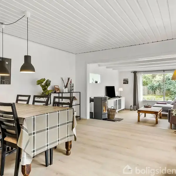 Spisebord dækket med ternet dug står i en lys stue med træstole og to sorte lamper hængende over. I baggrunden ses en åben opholdsstue med sofa og udsigt til haven.