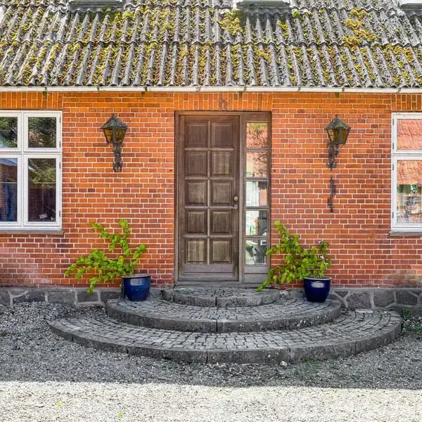 Rød murstensbygning med to vinduer og en central trædør. To potteplanter står på trappen op til døren. Muren er dekoreret med to væglamper.