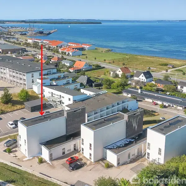 Bygning markeret med en rød pil ligger ved vandet. Dens moderne design består af hvide og grå facader. Omgivet af flere bygninger og parkeringsplads nær en kystlinie.