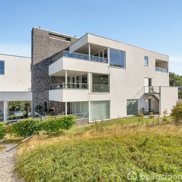 Moderne lejlighedskompleks står stilrent med glas- og betonstruktur. Store vinduer og altaner skaber lys og åbenhed. Det hele indrammes af grønne områder og en klar blå himmel.