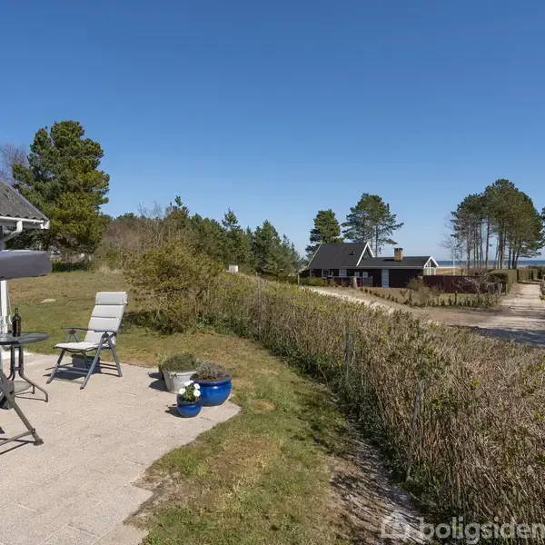 Gyngesofa og stol står på en terrasse, omkranset af buske og grønne områder. I baggrunden ses et hus og en grusvej, omgivet af træer under en klar blå himmel.