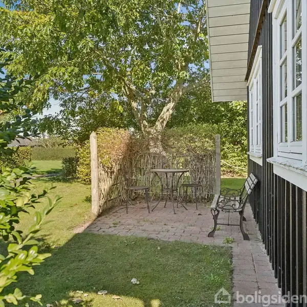 En lille terrasse med et rundt bord og to stole står på et flisebelagt område ved siden af et sort-hvidt hus. Til venstre er en grøn have med træer.