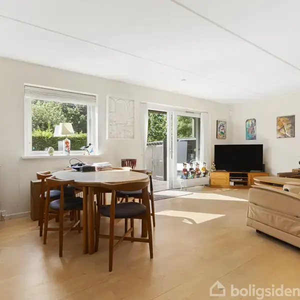 En stue med spisebord og stole står på et trægulv. I baggrunden er en sofagruppe og et fladskærms-tv. Stort vindue giver udsigt til en grøn have.