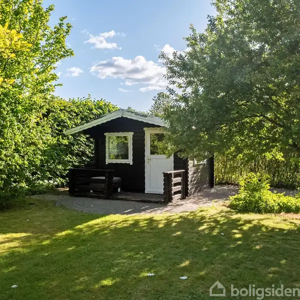Et sort-hvidt træhus står i en solrig have omgivet af grønne træer og buske. Huset har en lille terrasse foran med en bænk.