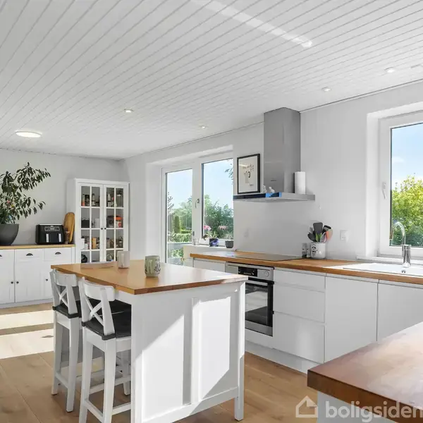 Køkken med hvidt interiør, to stole ved en ø, og træborde. Store vinduer giver udsigt til grønne træer udenfor; skabe og planter tilføjer indretning. Lys og moderne indretning.