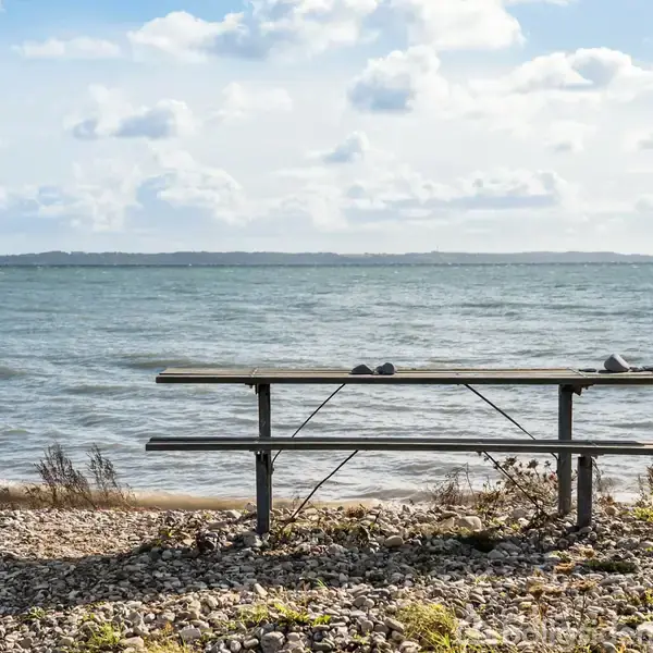 Et picnicbord står stille på en stenet strand bred ved en rolig sø, omgivet af spredt vegetation under en blå himmel med skyer.