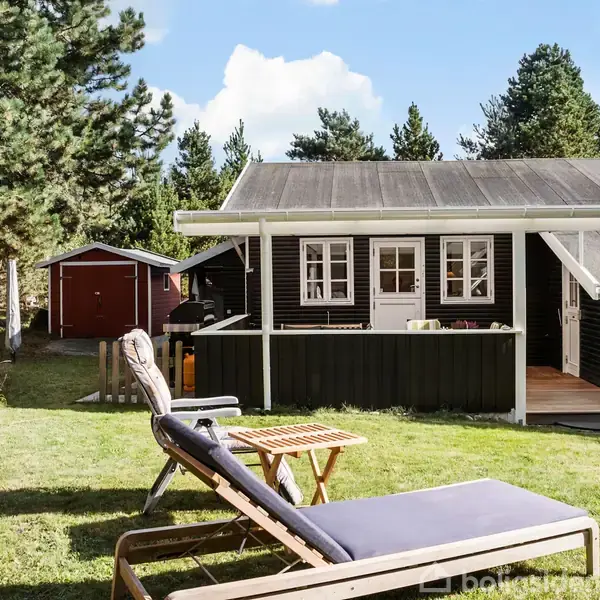 Et sort sommerhus står i en solrig have med en liggestol og et lille bord foran. I baggrunden ses flere træer og en rød skur.