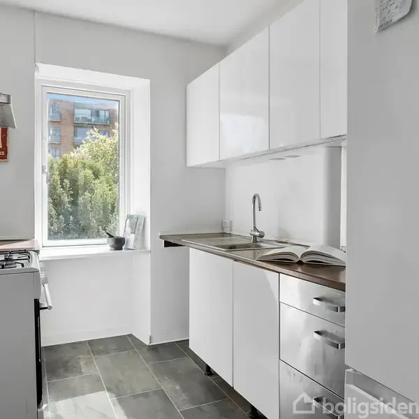Stue med sofa og puder står ved et vindue med åbent glasdør, der fører ud til en balkon. Blomsterpotter og små dekorationer pryder vindueskarmen. Lys træparketgulv.