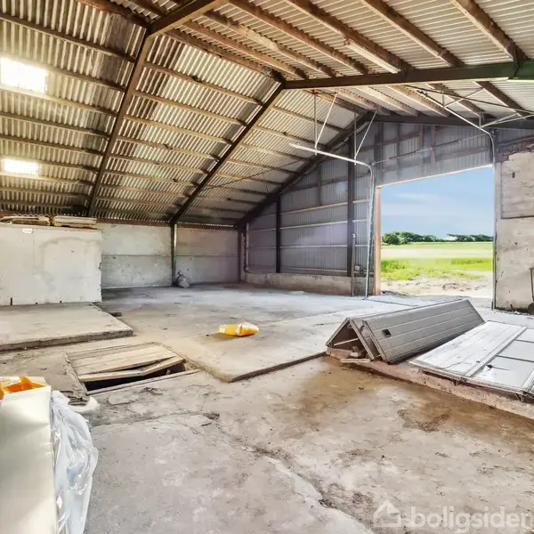 En rummelig stålhal med højt til loftet, åbner mod en mark. Indeni ligger byggematerialer og affald på betongulvet i en uindrettet og lys hal.