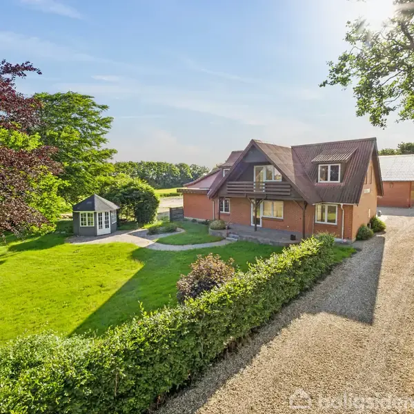 Et rødt murstenshus ligger i en stor, velplejet have med en grusvej og en pavillon. Træer omgiver ejendommen, skaber en fredfyldt, landlig atmosfære under en klar blå himmel.
