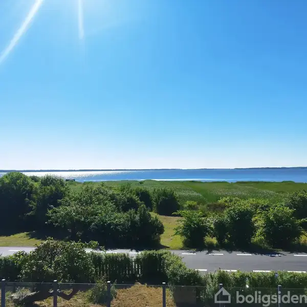 Vej strækker sig gennem grønt landskab med buske og træer. I baggrunden ses en stor sø under en lys blå himmel med sol.