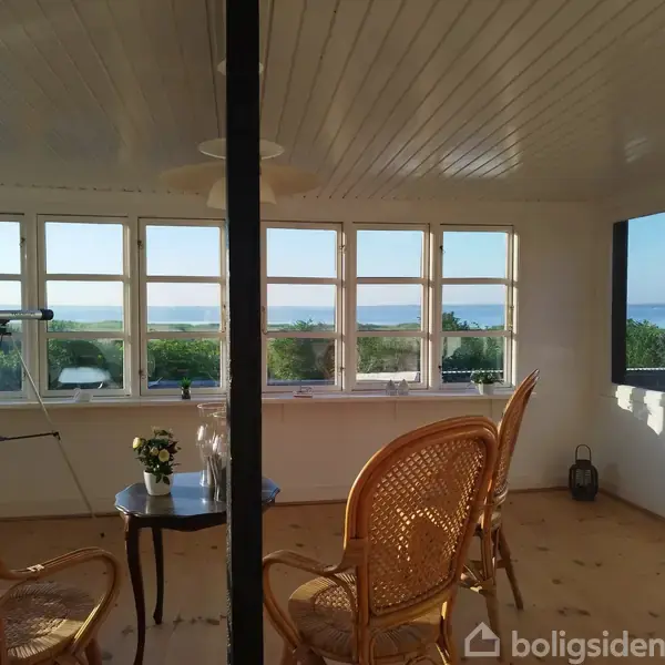 To rattan-stole står ved et bord med en vase i en lys udestue. Et teleskop er rettet ud mod et vindue med udsigt til havet og grønne buske.