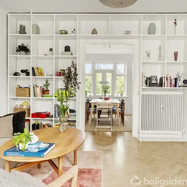 Reol fyldt med bøger og dekorationer står mod en hvid væg. Rundt sofabord og planter i forgrunden. Et tilstødende spiseområde med spisebord i baggrunden.