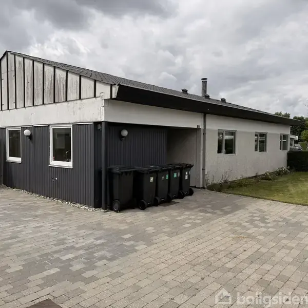 En enkeltstående, moderne bungalow med fladt tag, bemalet i hvide og grå nuancer. Skraldespande står langs væggen. Beliggende på en brolagt gårdsplads med græs og buske i baggrunden.