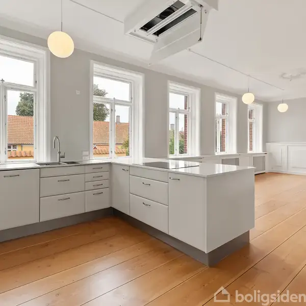 Køkken i åben plan med hvidt interiør og stor køkkenø. Store vinduer giver lys, mens træbjælker og moderne lamper skaber elegance. Trægulv og udsigt til hustage fuldender rummet.