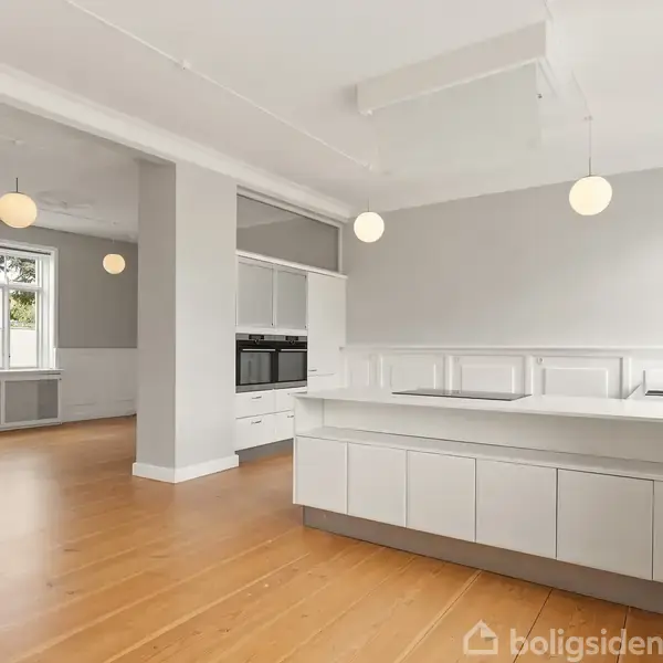 Et lyst, moderne køkken med en stor køkkenø og integrerede ovne. Køkkenet er i et åbent rum med trægulve og hvide vægge, omkranset af store vinduer med udsigt til grønne områder.