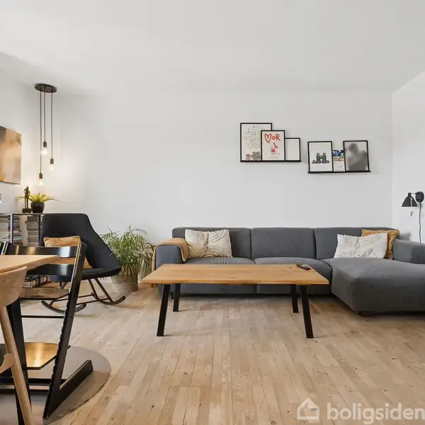 En grå sofa står i en lys stue, dekoreret med hvide vægge, træmøbler og væghængte billeder. Et spisebord med stole er placeret til venstre, nær en glasdør.