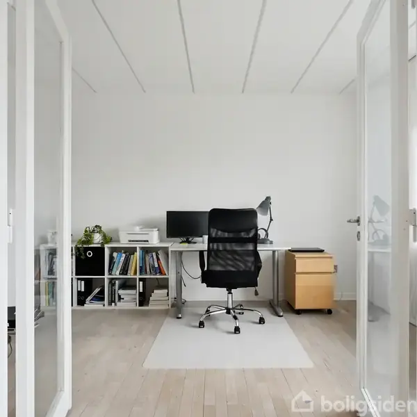 Et skrivebord med en computer og lampe står i et lyst kontorrum. En sort kontorstol er foran skrivebordet. En bogreol ved siden af har bøger og planter.