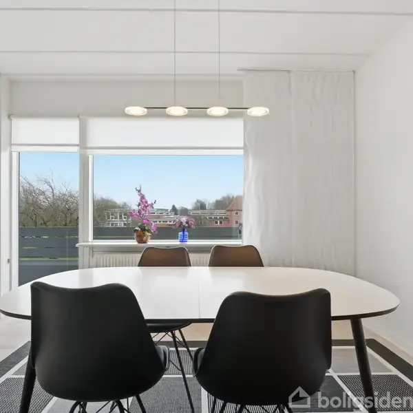 Et ovalt bord med sorte stole står i en lys spisestue. Bordet er dækket med en vase med blomster og en flaske. Store vinduer giver udsigt til et træfyldt område.