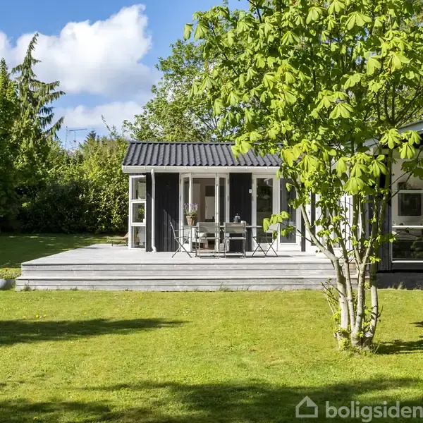 Et sommerhus med sort træbeklædning står på en velplejet græsplæne. På terrassen er der møbler til afslapning. Omgivet af træer under en blå himmel med hvide skyer.