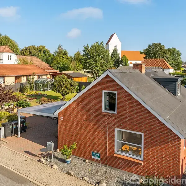 Rødt murstenshus med sort tag står i en pæn forhave med buske og blomster. Ved siden af er en carport. Omkringliggende huse og træer i en rolig villavej.