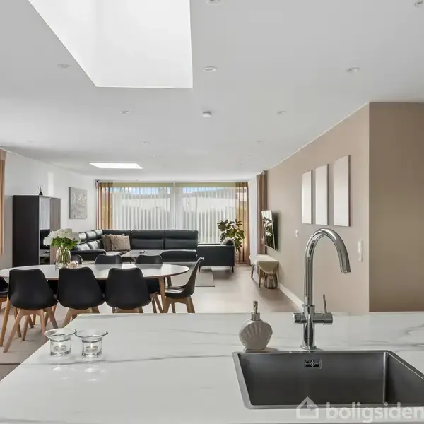 Køkken med marmorvask, bordplade og indbygget armatur. Foran bordet står en sort spisebordsstol, og en stue med sofa og store vinduer ses i baggrunden. Ingen tekst bortset fra "Boligsiden".