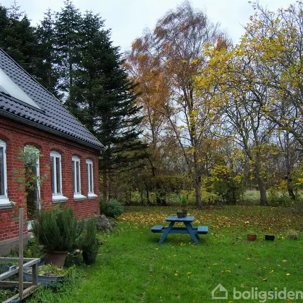 Rødt murstenshus med hvide vinduer står ved en frodig have. En blå bænk står på en grøn græsplæne, omgivet af træer med efterårsblade.
