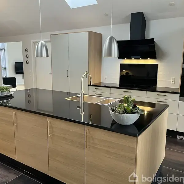 Køkkenø med sort bordplade og indbygget vask; stående i et moderne køkkenmiljø med indbygningslamper, grå skabe, ovne og komfur. To hængende metalpendler.