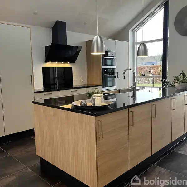 Køkkenø med træskabe og sort bordplade står centralt. En lampe hænger over øen. Bagved ses en væg med ovne, skabe og en emhætte. Vindue til højre med udsigt.