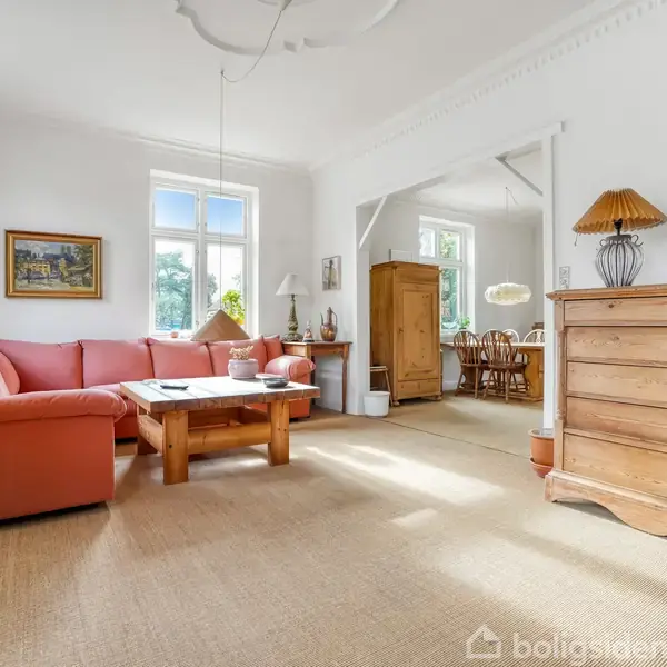 Rød sofa står stille ved et træbord i en lys stue med store vinduer. En kommode med lampe, maleri på væggen, og en spiseafdeling i baggrunden.