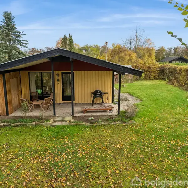 Et lille træhus med en overdækket terrasse har havemøbler og en grill. Det står på en græsplæne omgivet af træer og buske under en blå himmel.