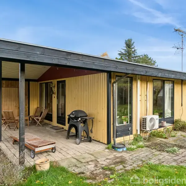 Et gulmalet træhus med sort tag og store vinduer står på en havegrund. Der er en overdækket terrasse med havemøbler og en grill. En græsplæne omgiver området.