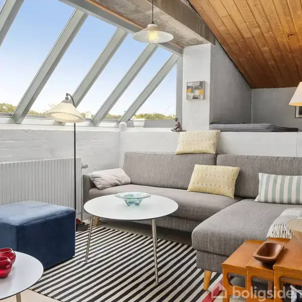 Grå sofa med farvede puder står i en bolig med skråvinduer og trægulv. Rundt sofabord, stribet tæppe, blå skammel, og brunt sidebord med lamper giver stuen et hyggeligt udtryk.