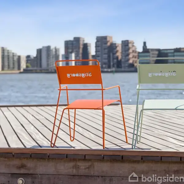 To farvede stole står på en træterrasse ved en havn, med vand og byens skyline i baggrunden. Stolenes ryglæn har teksten "Boligsiden".