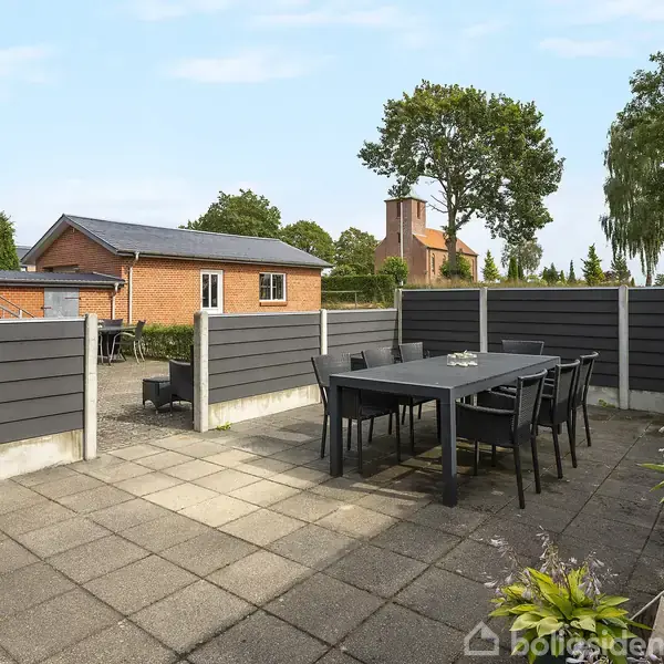 Udendørs terrasse med sort spisebord og stole, beliggende på flisebelagt område. Parasol står sammenfoldet ved siden. Omkranset af grå hegn. Rød teglstensbygning og træer i baggrunden.