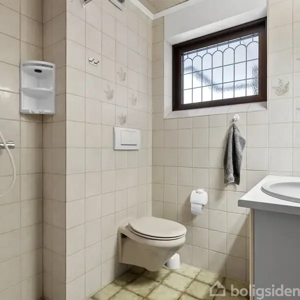 Toilet hænger på væggen, ved siden af en håndvask med spejl i et flisebeklædt badeværelse. Bruser til venstre, håndklæde hænger ved siden af et vindue.