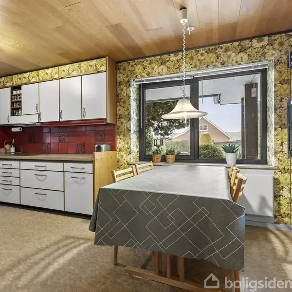 Køkken med et spisebord dækket af dug. Skabe og skuffer i retrostil mod en rød flisevæg. Vinduesudsigt til have, og en loftslampe hænger over bordet.