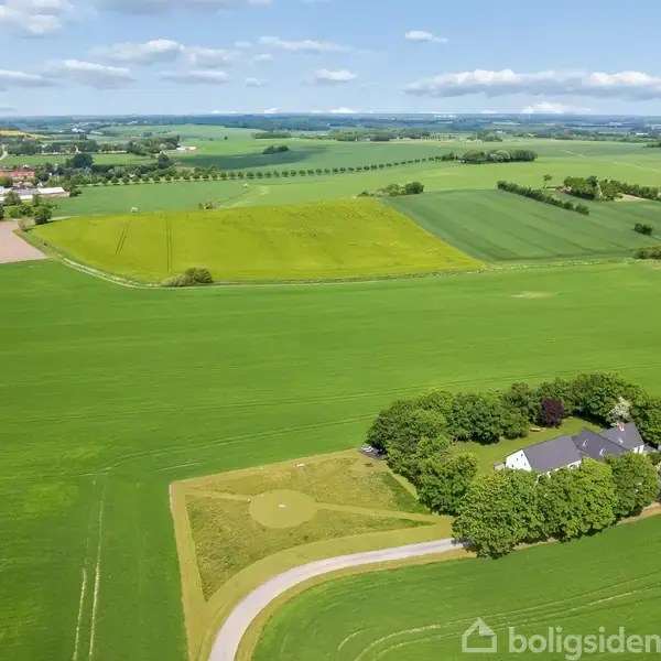 En stor gård omgivet af tætte træer ligger midt i et grønt landskab med marker, under en blå himmel med spredte skyer. En snoet vej fører til ejendommen.