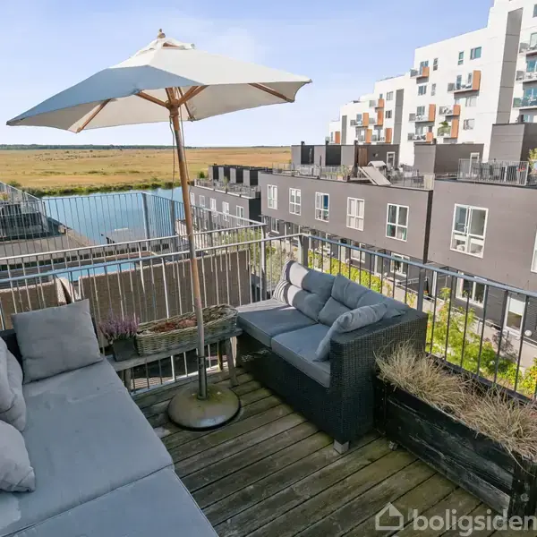 Terrasse med grå sofaer og parasol; placeret på trægulv. Omgivet af moderne, grå lejlighedsbygninger og udsigt over åbne marker. God plads til afslapning og udendørs ophold.