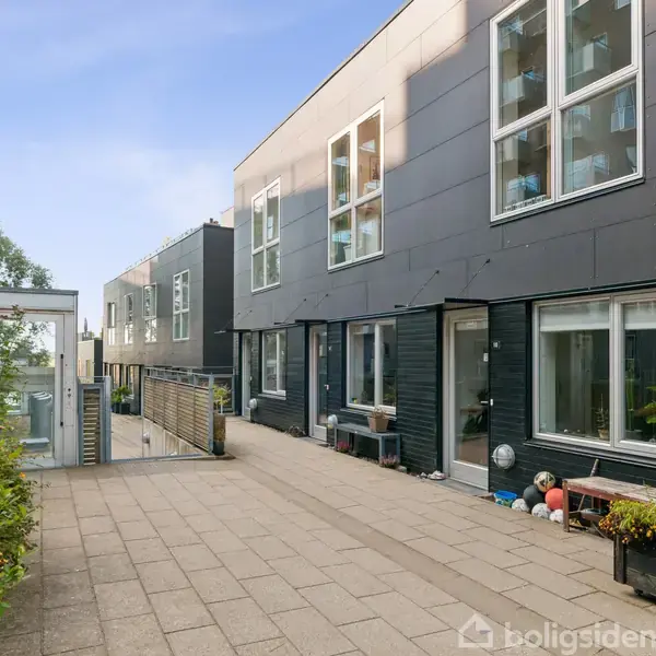 Bygning med sorte facader og store, hvide vinduer står på en flisebelagt gangsti med plantekasser. Omkringliggende bygninger og træer er synlige i en urban indstilling.