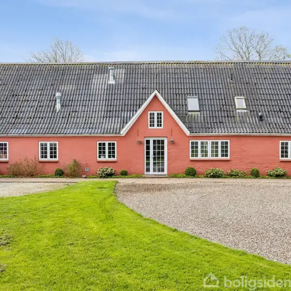 En lang, lyserød landejendom med sorte teglsten på taget. Bygningen har flere vinduer og en hvid hoveddør i midten. Foran er der en grusvej og græsplæne.